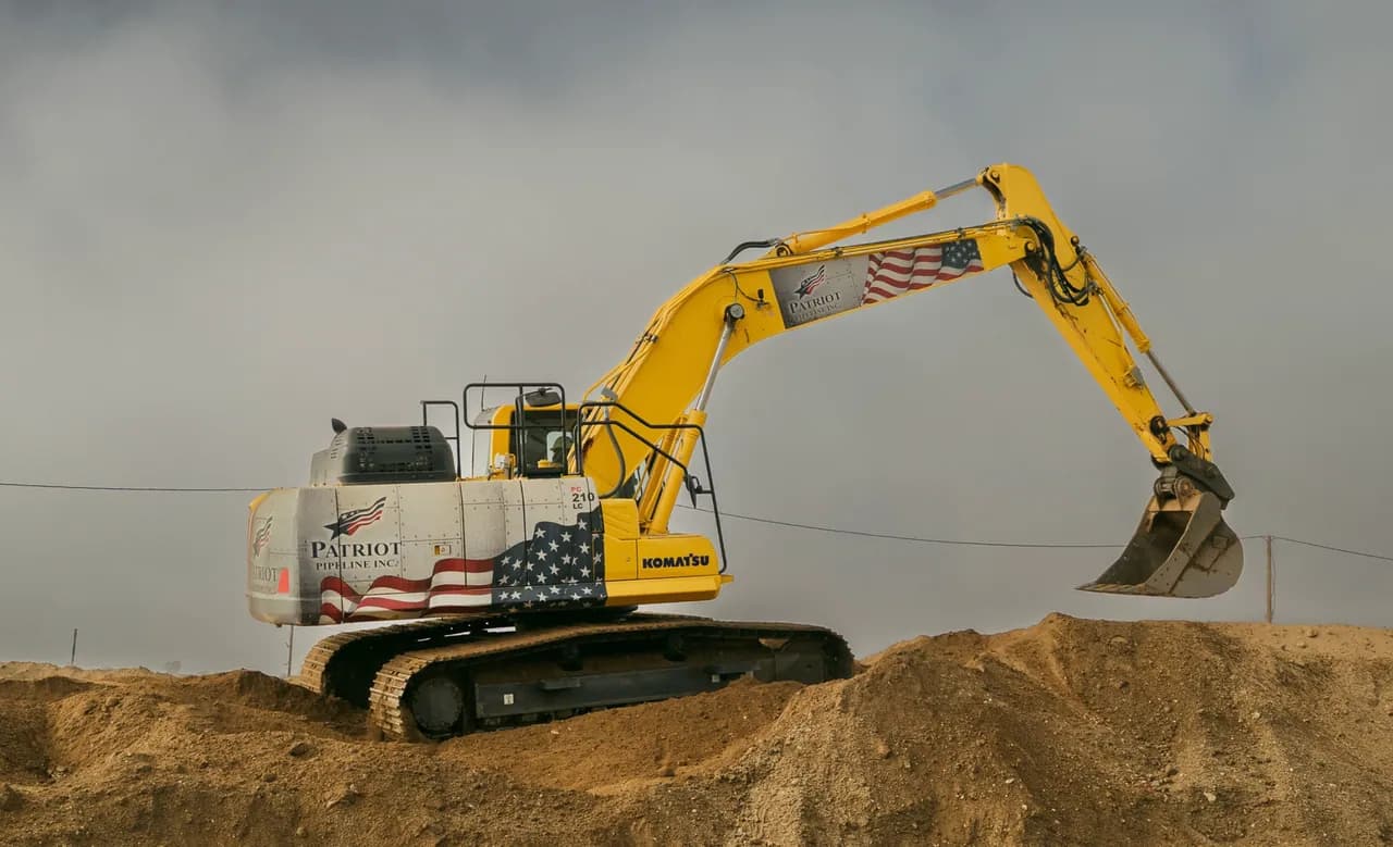 Patriot Pipeline Komatsu excavator in flag livery, bucket raised over a dirt stockpile